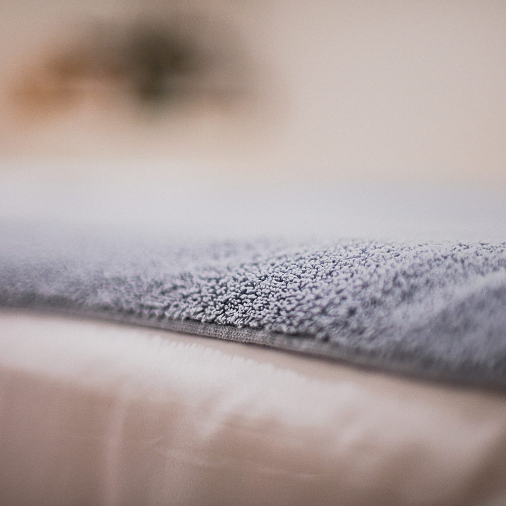 A close view of the folded edge of a soft grey-blue towel resting on white linen, its looped pile sharp in the foreground and the studio falling away out of focus behind.