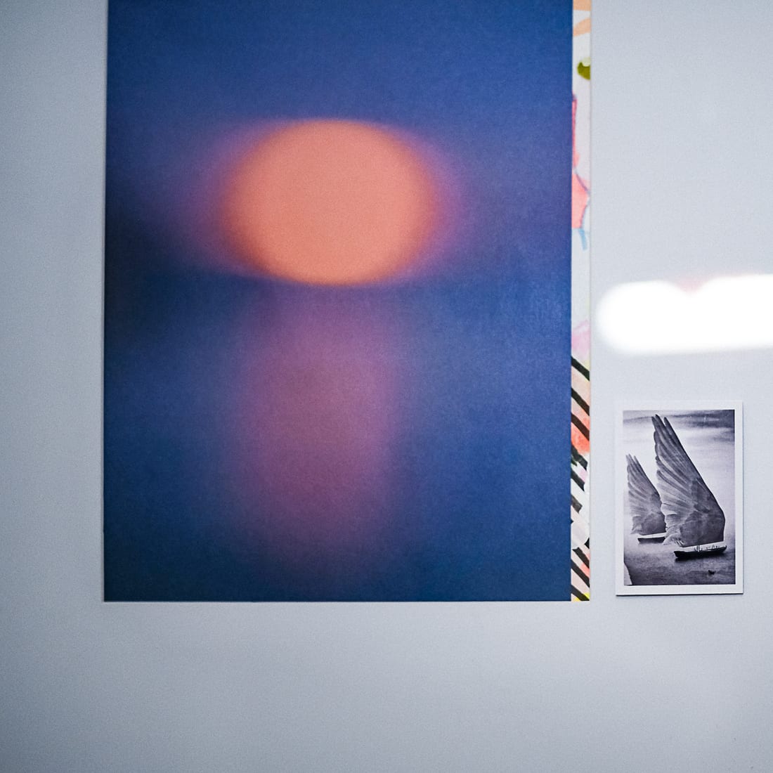 A corner of the studio: a large print of an orange sun over deep blue, and beside it a small black-and-white photograph of a boat whose sails are a pair of outstretched wings.
