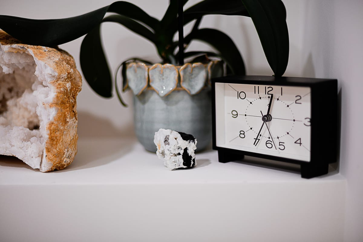 A shelf in the studio: a large open geode, a small black-and-white crystal, an orchid in a pale ceramic pot, and a plain black desk clock.
